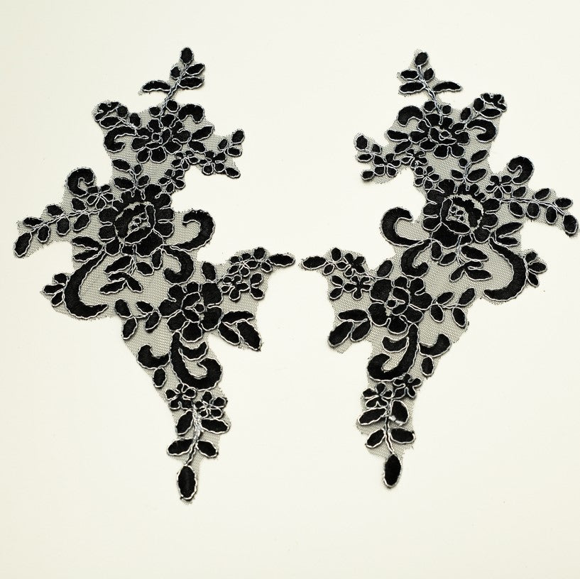 Black embroidered applique pair edged with silver metallic silver cord laying flat on a white background.