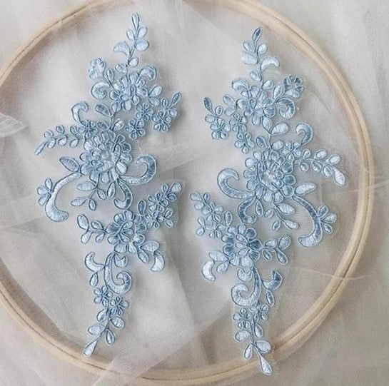Mirrored pair of baby blue corded floral lace appliques.  The appliques are laying flat on white netting inside a wooden embroidery ring.       