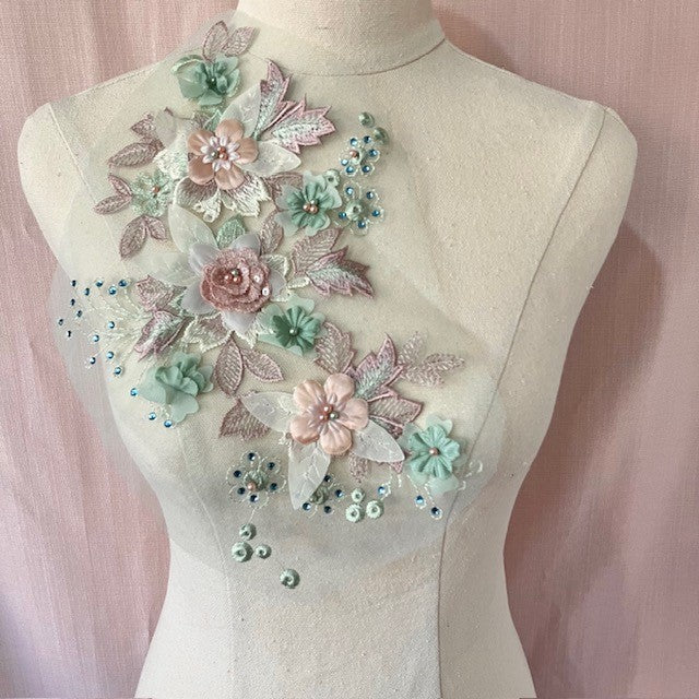 A large 3d floral applique in dusty pink and sage green. The flowers have pearl centres . Blue gems highlight the leaf shapes. Displayed on a mannequin.