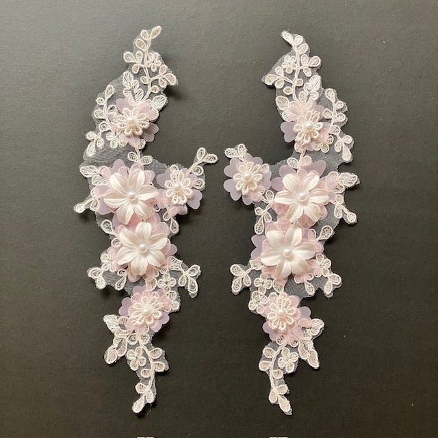 A white corded floral applique pair with 3d white flowers . Each flower has a pearl centre .A layer of pink chiffon forms the petal shape at the base of each flower. 
