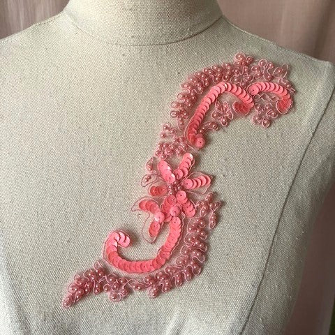 An applique with coral sequin strings formed into an S  with a star shape in the centre . The S is surrounded by tiny coral pearl bubbles .