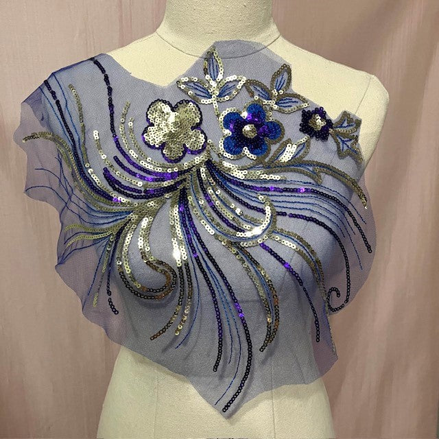A large royal blue applique with a 2D gold sequinned flower and a 2D blue sequinned flower surrounded by sprays of gold and purple sequin  stems in a swirled design on a blue net backing. 