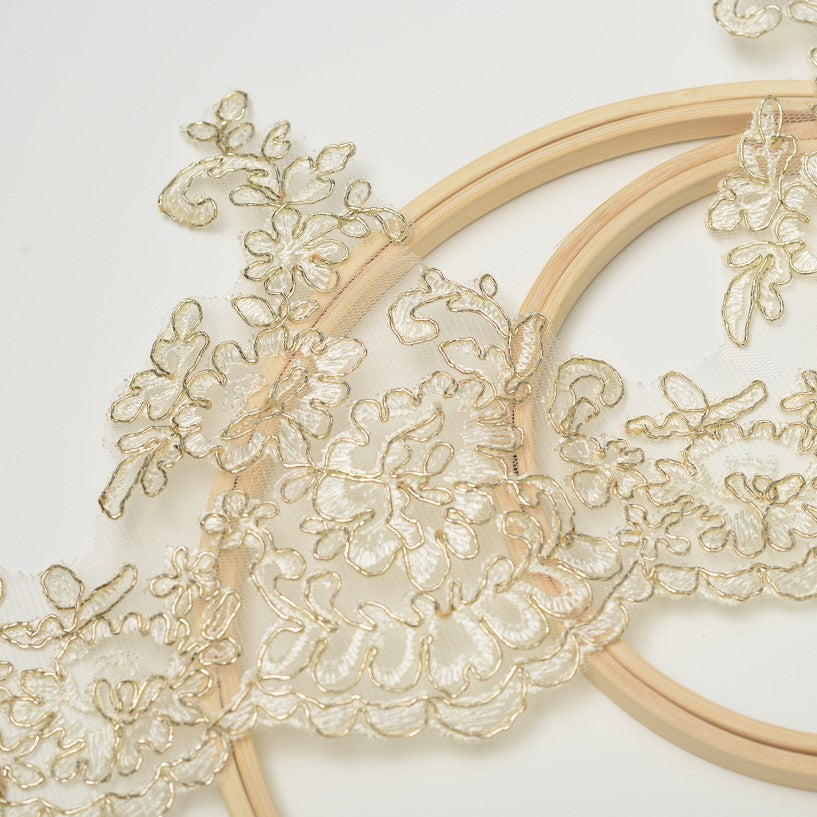 Gold corded cream lace border with a floral design and a scalloped lower edge laying diagonally across wooden embroidery rings..