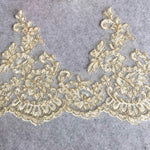 Gold corded cream lace border with a floral design and a scalloped lower edge laying flat on a mottled grey background.