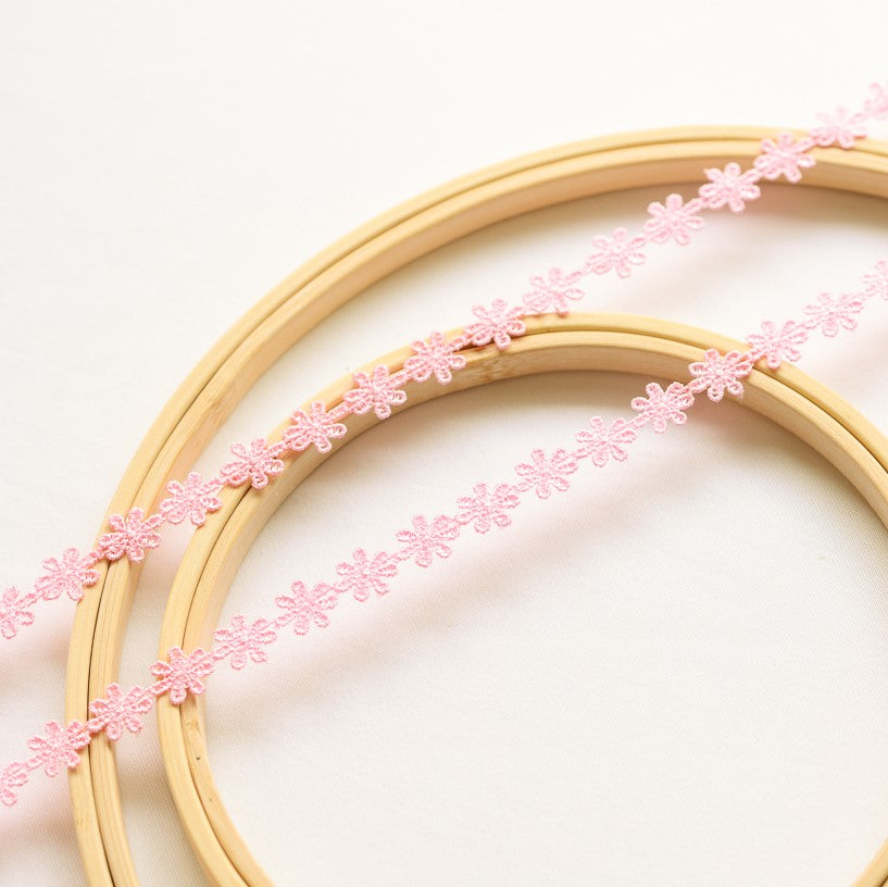 Two strands of delicate medium pink daisy lace laying flat across wooden embroidery frames on a white background.