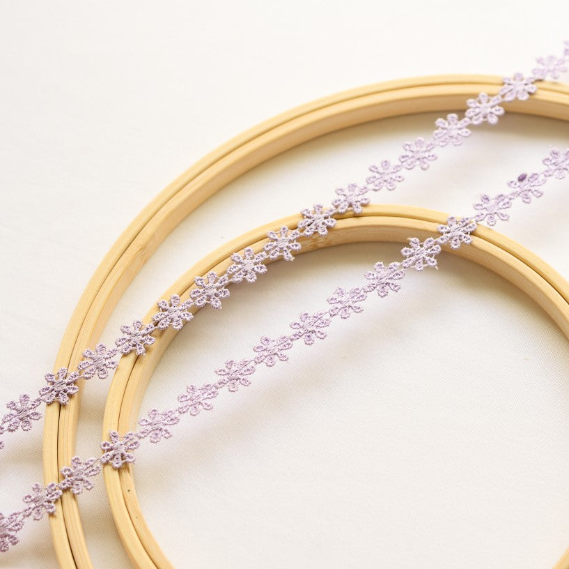 Two strands of delicate mauve daisy lace laying flat across wooden embroidery frames on a white background.