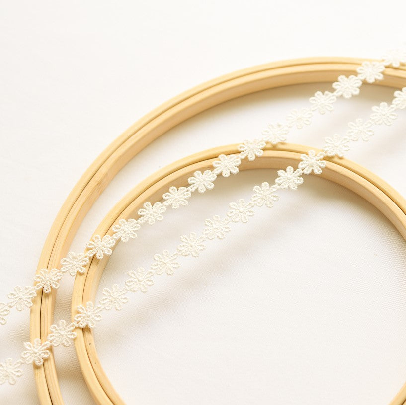 Two strands of delicate off-white daisy lace laying flat across wooden embroidery frames on a white background.