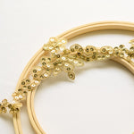 A narrow gold heavily sequinned trim in a  scalloped tiara shaped pattern laying diagonally across wooden embroidery hoops on a white background.  The trim is edged with gold cord.