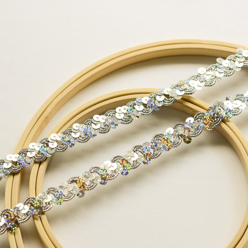 A silver sequinned trim in a wave design . The silver laser hologram sequins reflect blues and greens.  Two strands of the trim lay across wooden embroidery rings on a white background.