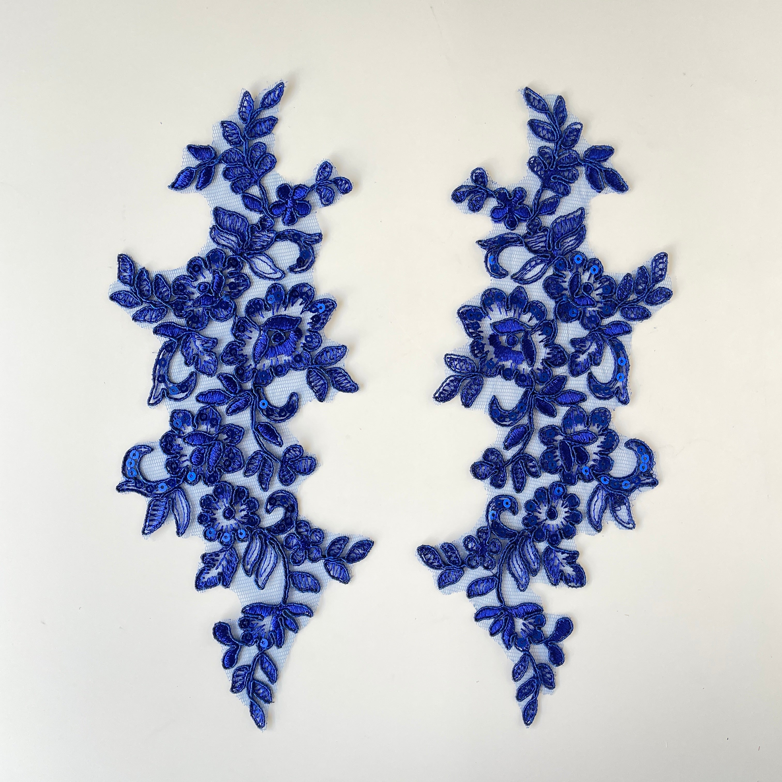 Mirrored pair of royal blue appliques sprinkled with royal blue sequins on the petals in the floral design.  The appliques are laying flat on a white background.