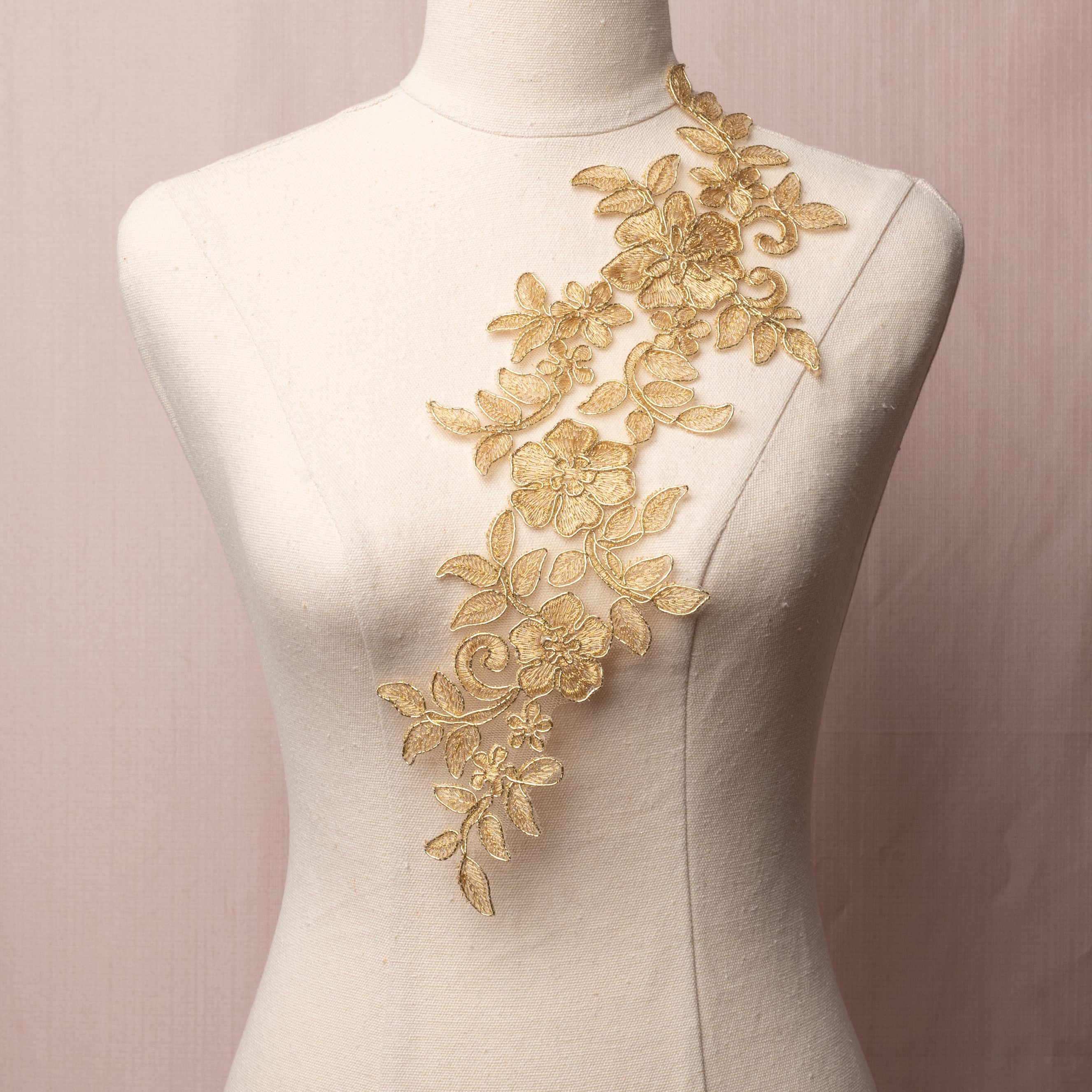 Single trailing floral applique embroidered with metallic gold thread onto a net backing and edged with metallic gold cord.  The applique is displayed on a mannequin.