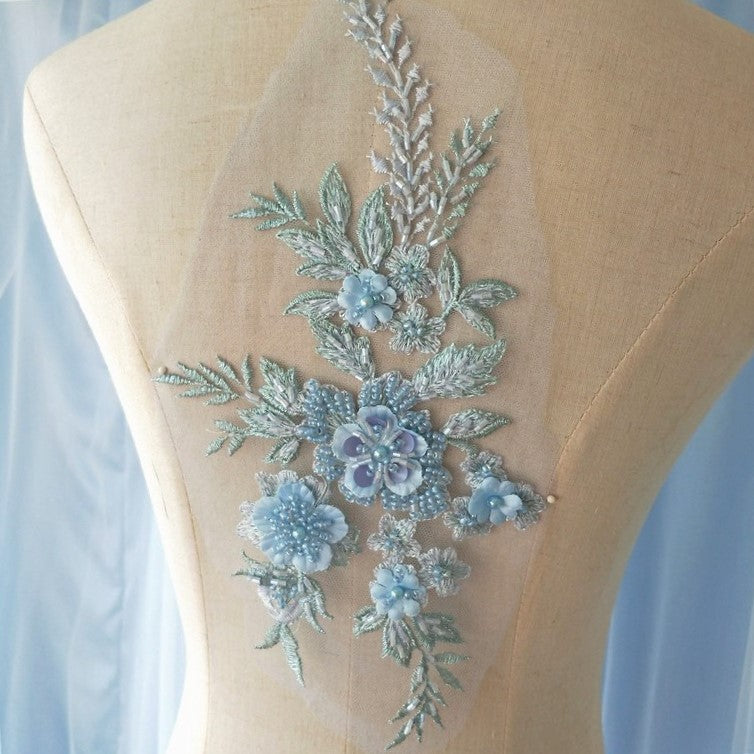 Heavily beaded single blue floral applique embroidered with a blue grey metallic thread and embellished with blue 3D flower petals. The applique is displayed on a mannequin.