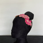 Fuchsia headpiece with a central 3D flower.  The headpiece is edged with gold cord and embellished with AB crystals and displayed on a mannequin..