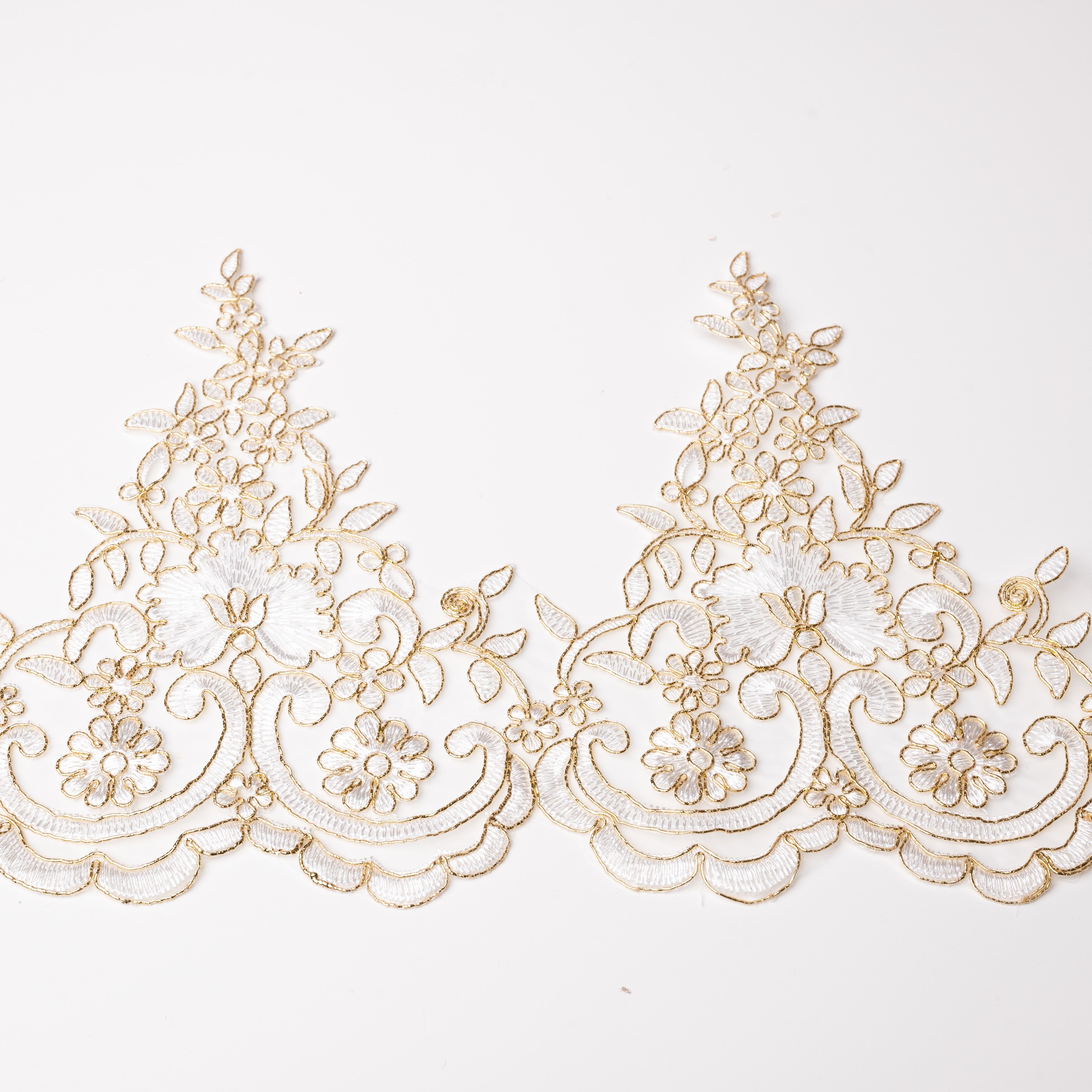 White embroidered lace border with a scalloped lower edge.  The floral pattern is edged with a fine gold metallic cord giving the lace an overall gold shimmer.  The lace is laying flat on a white background.