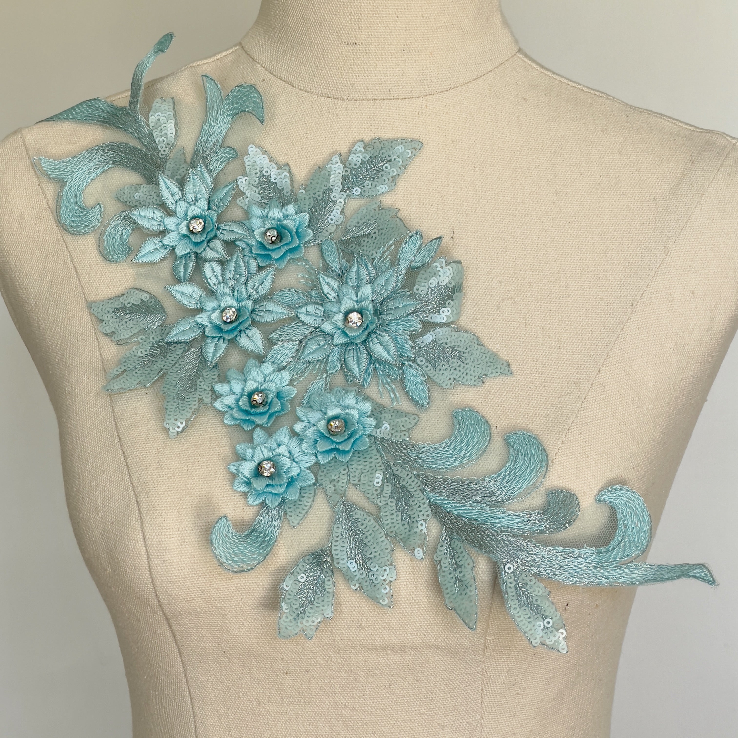 Single light blue floral applique embroidered with metallic thread and embellished with sequinned leaves and 3D flowers that have a crystal centre.  The applique is displayed on a mannequin. 