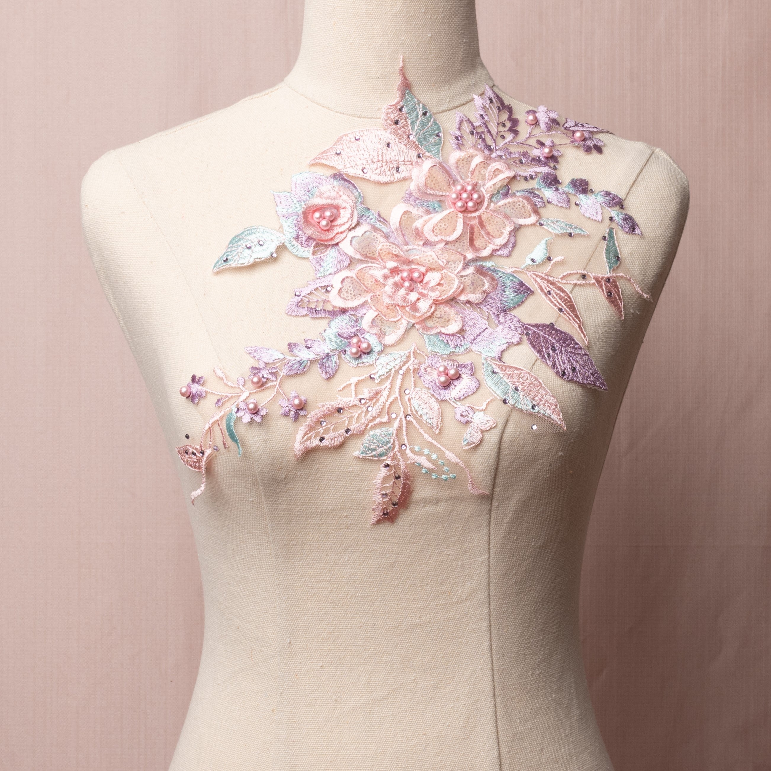 Single 3D floral applique in pastel shades of pink, lavender and green.  The 3D flowers have pearl centers and sequinned petals.  The applique is sprinkled with purple gems and is displayed on a mannequin.