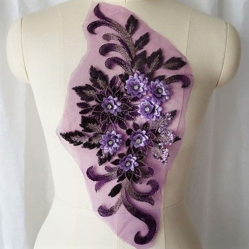 Single purple floral applique embroidered with metallic thread and embellished with sequinned leaves and 3D flowers that have a crystal centre.  The applique is displayed on a mannequin. 