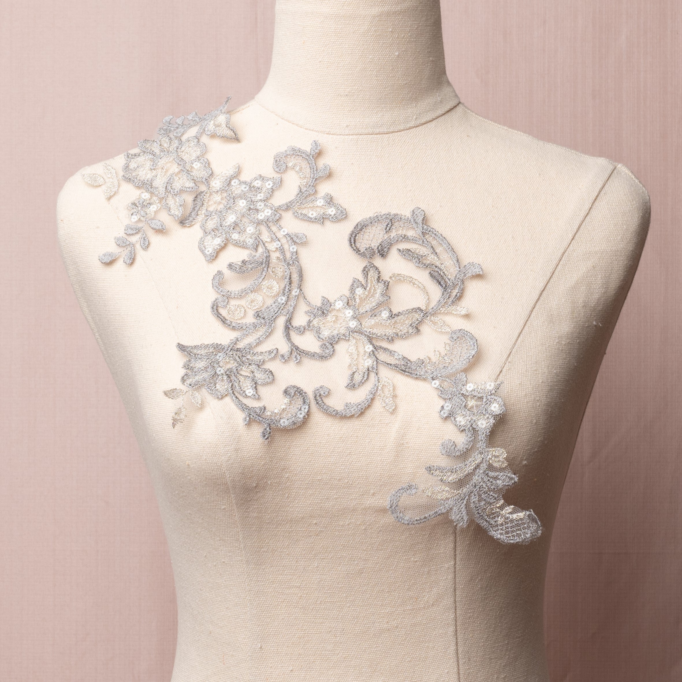 A single delicate grey floral applique highlighted with silver thread on the leaves and flower petals.  The applique is embroidered onto a very fine net and sprinkled with clear sequins.   The applique is displayed on a mannequin.