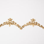 A narrow gold heavily sequinned trim in a  scalloped tiara shaped pattern laying on a white background.  The trim is edged with gold cord.