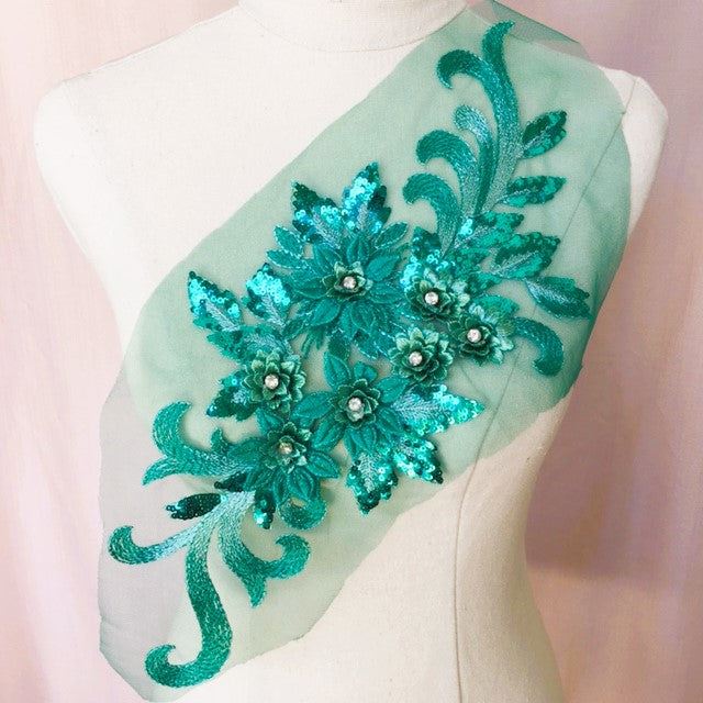 Single turquoise floral applique embroidered with metallic thread and embellished with sequinned leaves and 3D flowers that have a crystal centre.  The applique is displayed on a mannequin. 