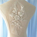 The applique's floral design is embroidered onto a fine net with a ivory white polyester thread and beaded with various styles of tiny beads. Small ivory bugle beads backed with clear eye shaped beads highlight the veins and stems of the leaf shapes and leaf sprays .Rows of translucent rondelle beads cover the outer embroidered petals and the  white 3D petals . Clear sequins overlay the white petals of the central flower . A larger ivory pearl forms the flower centres .