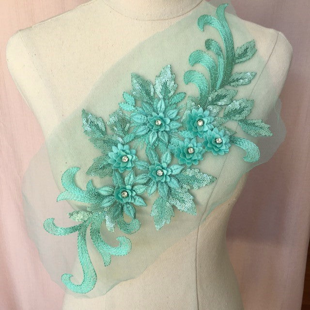 Single aqua green floral applique embroidered with metallic thread and embellished with sequinned leaves and 3D flowers that have a crystal centre.  The applique is displayed on a mannequin. 