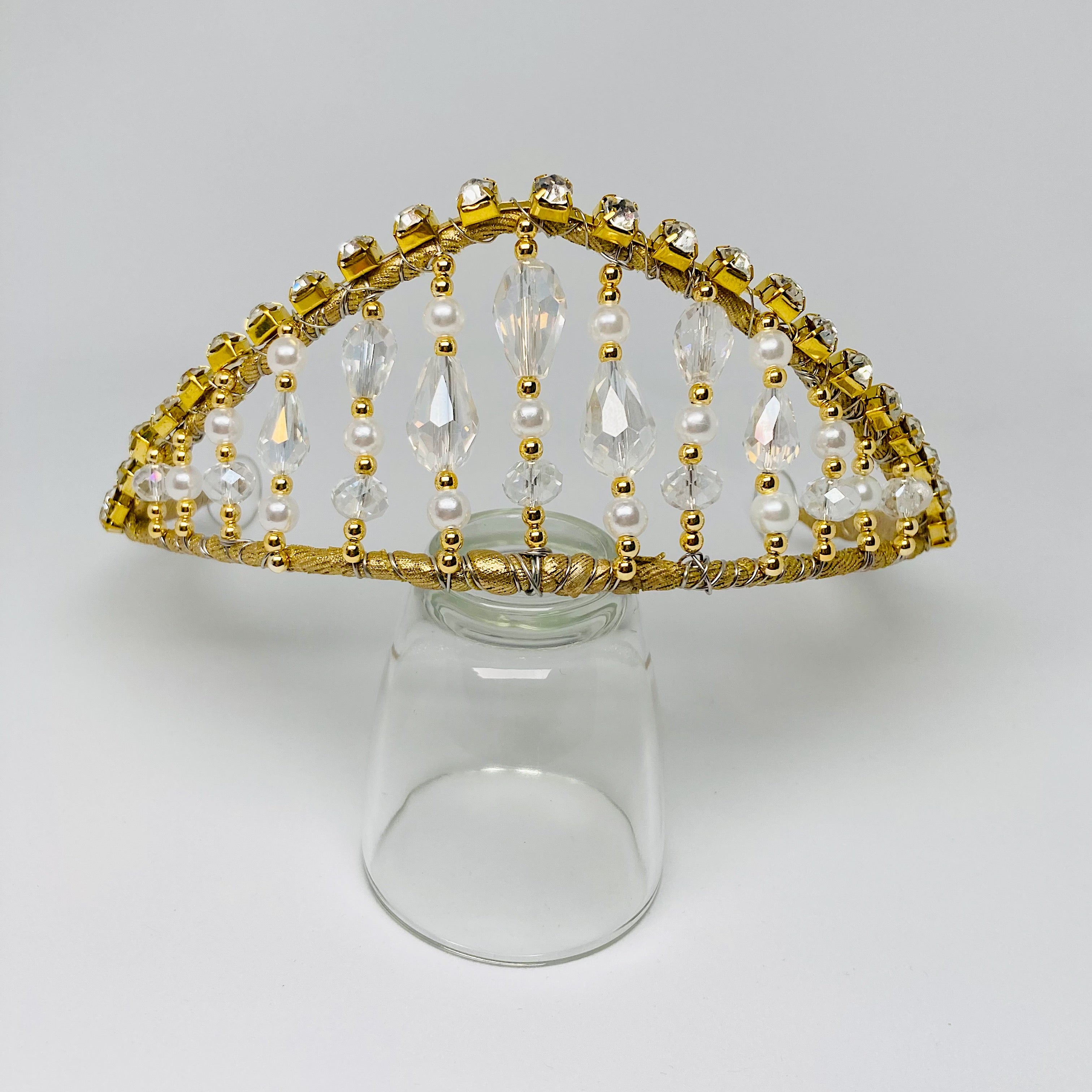 Front view of gold tiara embellished with rhinestones, clear crystals, gold beads and pearls.  The tiara is resting on a clear glass. 