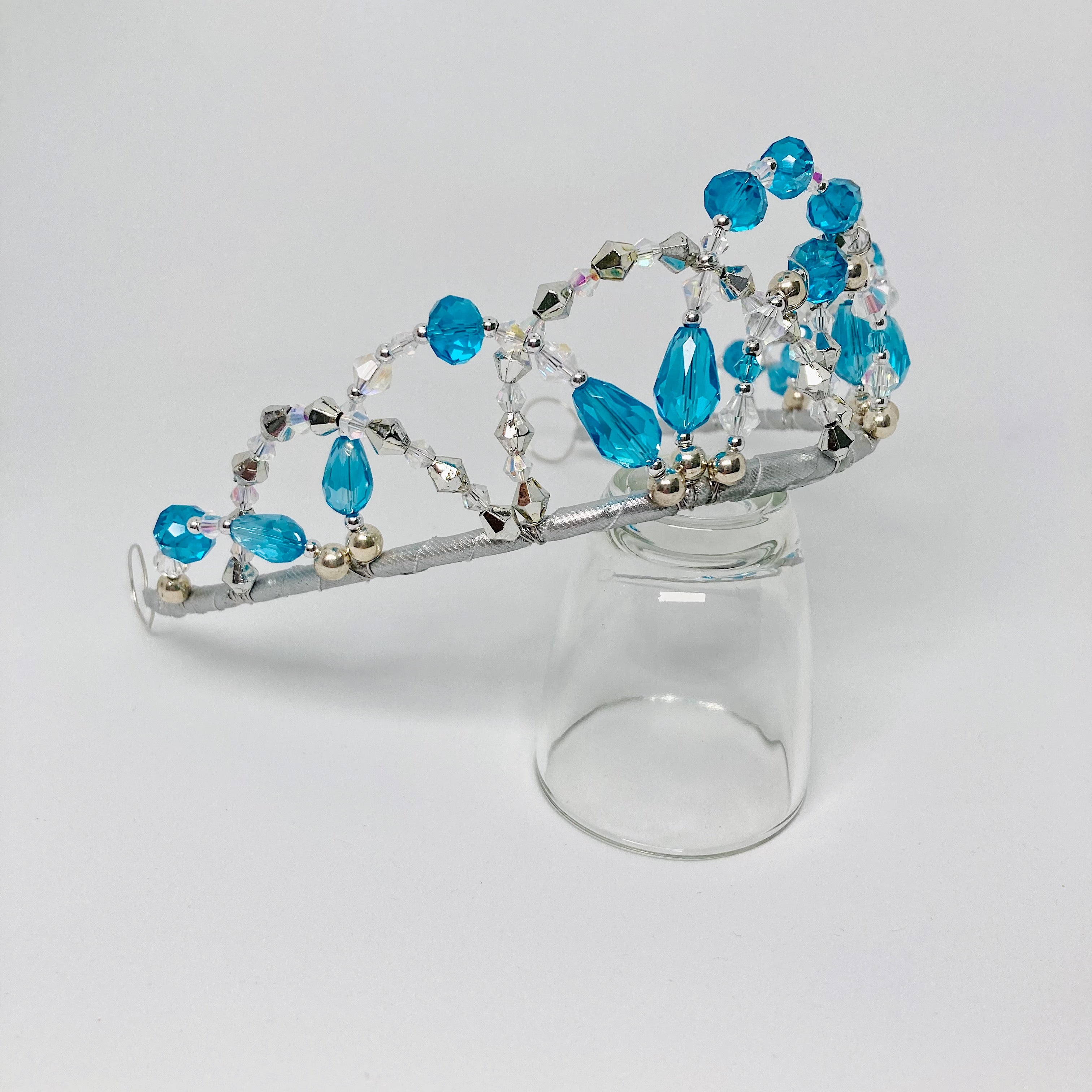 Side view of tiara featuring aqua blue and clear crystals and silver bicone and round beads.  Tiara is displayed on a clear glass.