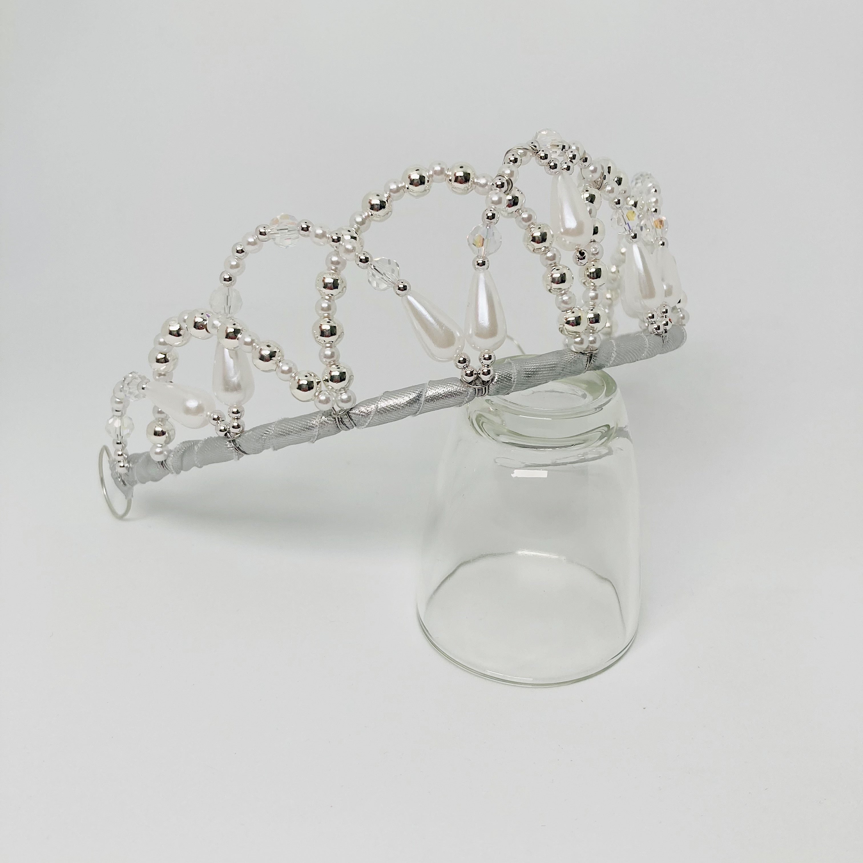 Side view of a tiara embellished with clear crystal, silver and frosted white beads.  The tiara is resting on a clear glass. 