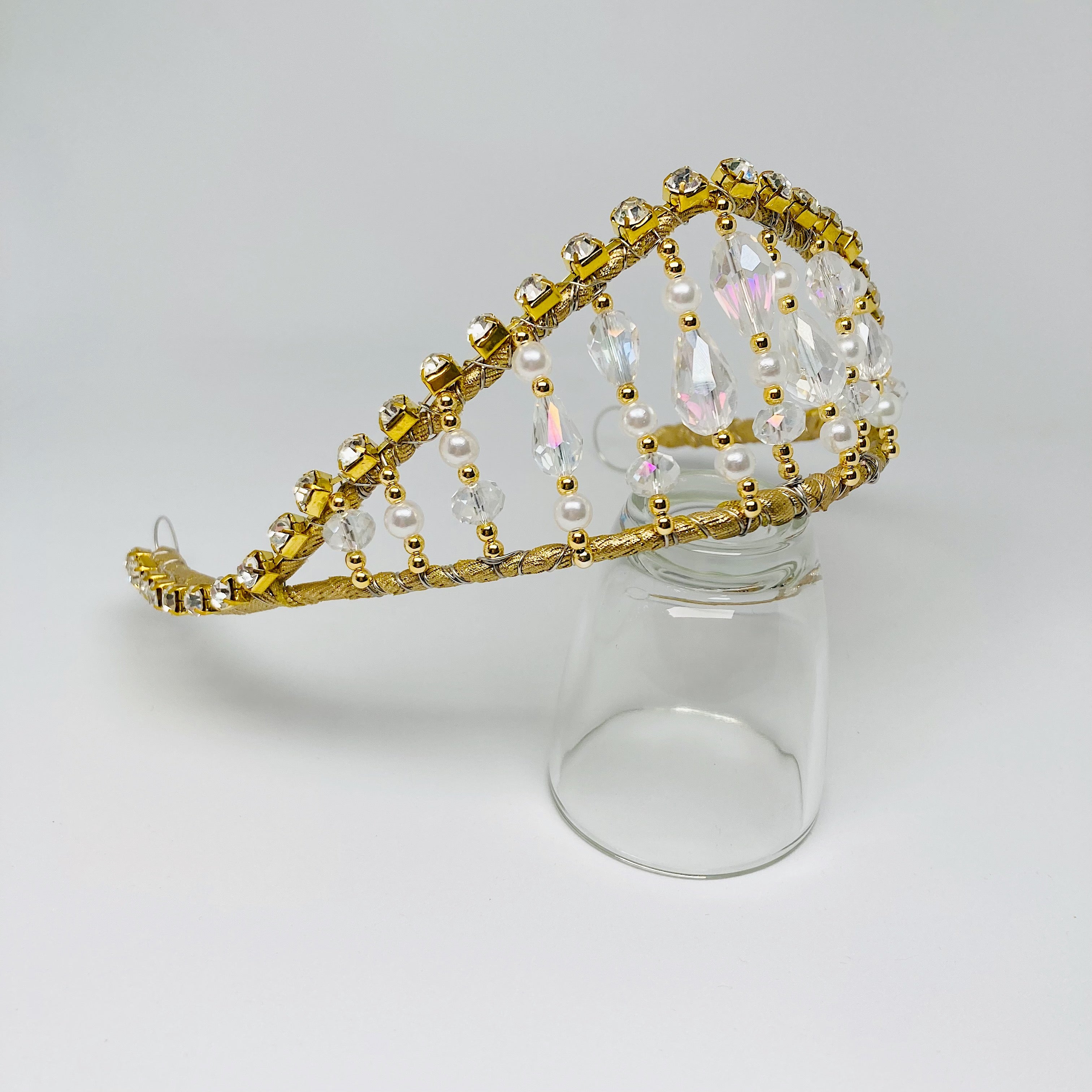 Side view of gold tiara embellished with rhinestones, clear crystals, gold beads and pearls.  The tiara is resting on a clear glass. 
