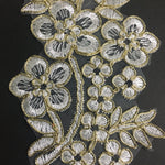 Close up view of white floral applique embroidered onto an organza background.  The flower, leaf and stem shapes are edged with gold cord.