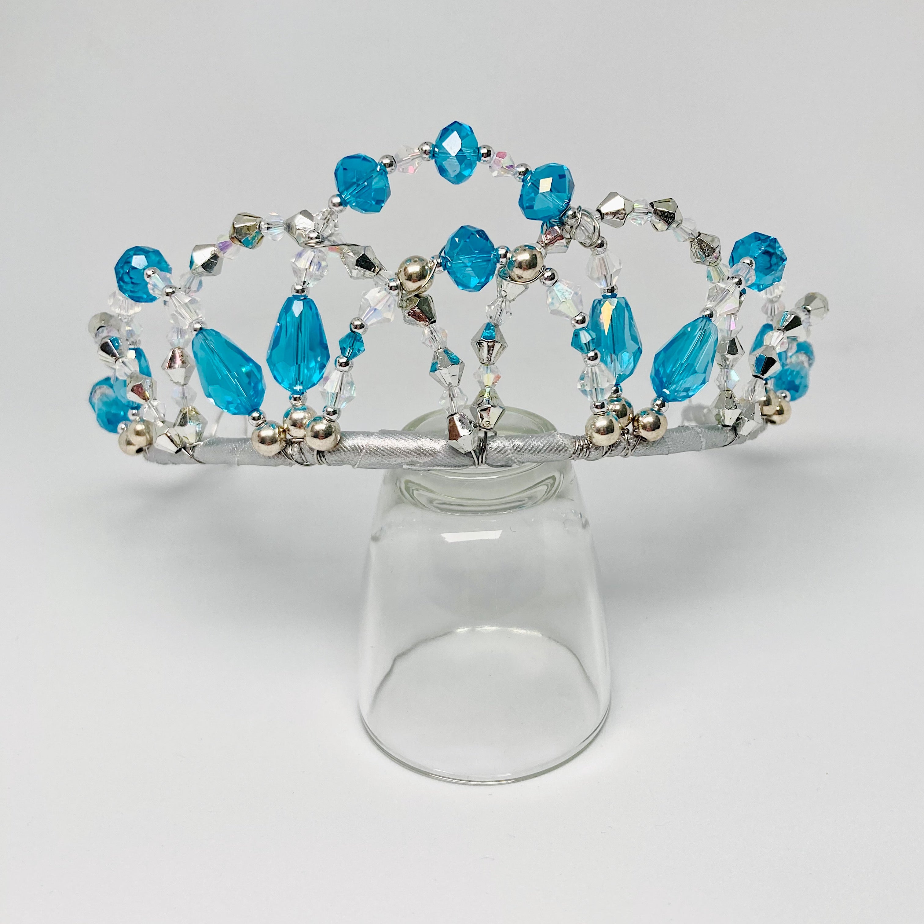 Tiara featuring aqua blue and clear crystals and silver bicone and round beads.  Tiara is displayed on a clear glass.