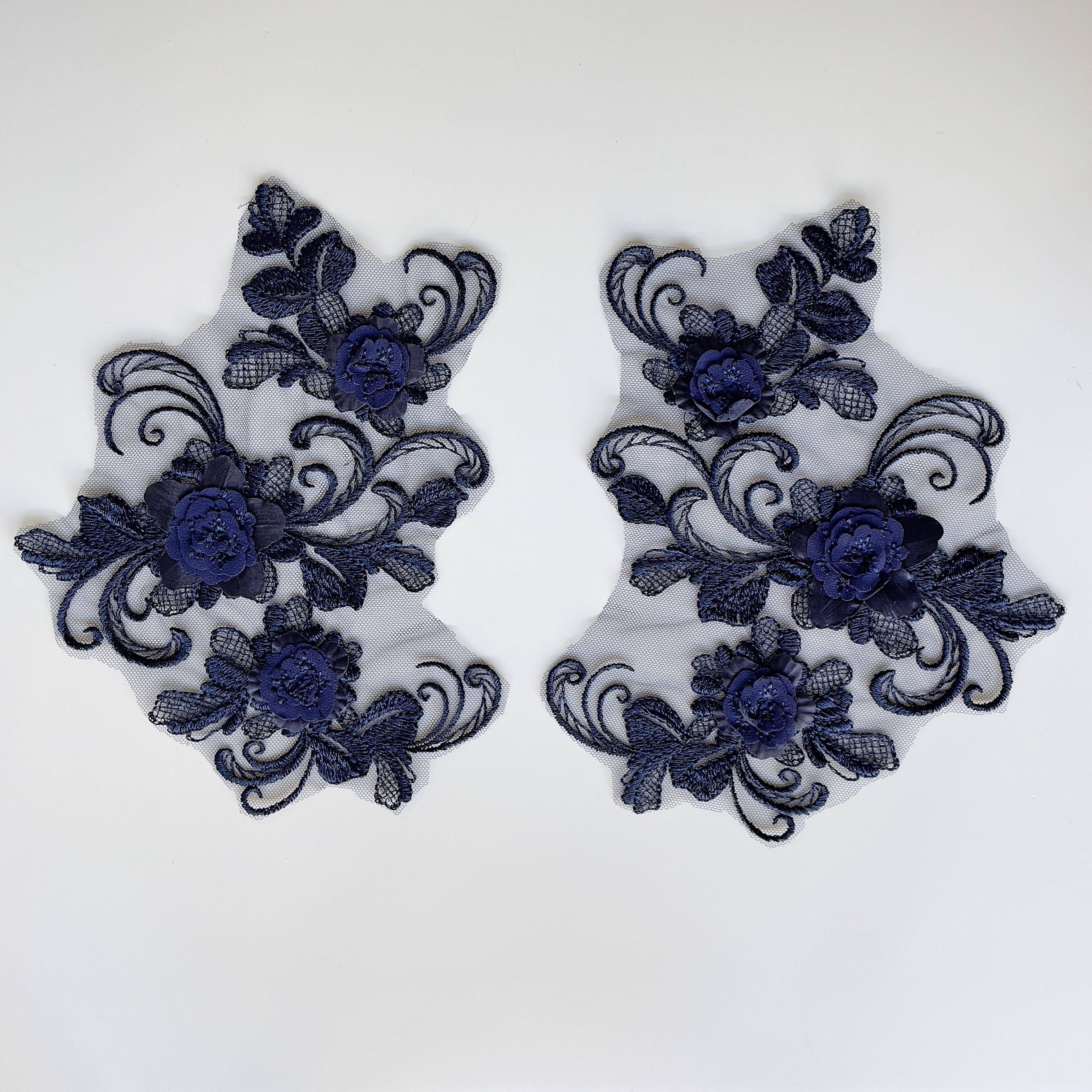 An openwork embroidered black applique pair in a floral design with swirling stems, 3D flowers and crosshatched leaves , laying flat on a white background.