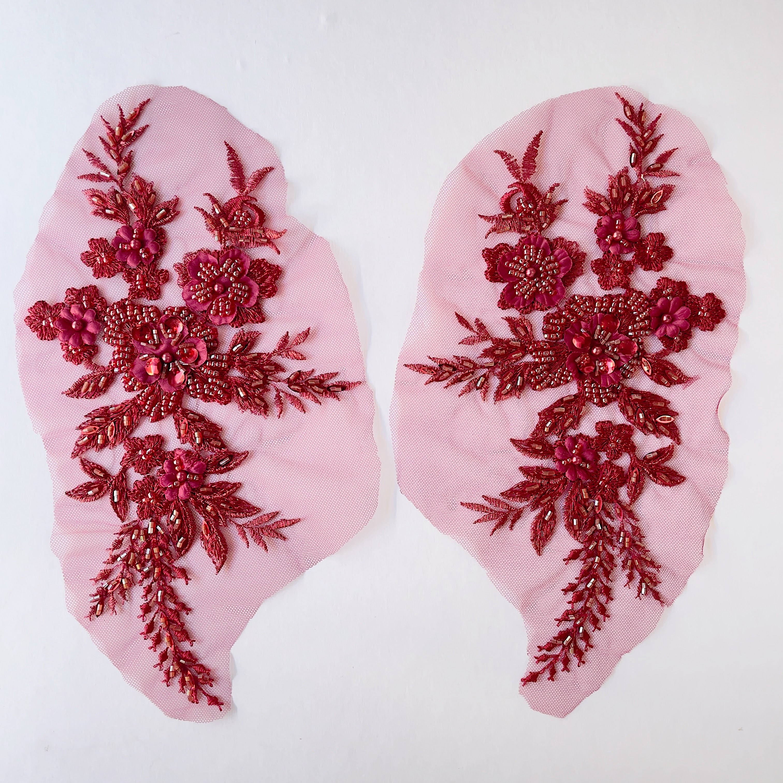 A mirrored pair of beaded burgundy floral appliques embroidered onto a matching net backing. Embellished with burgundy beads, sequins and satin 3D flowers.  The appliques are laying flat on a white background.