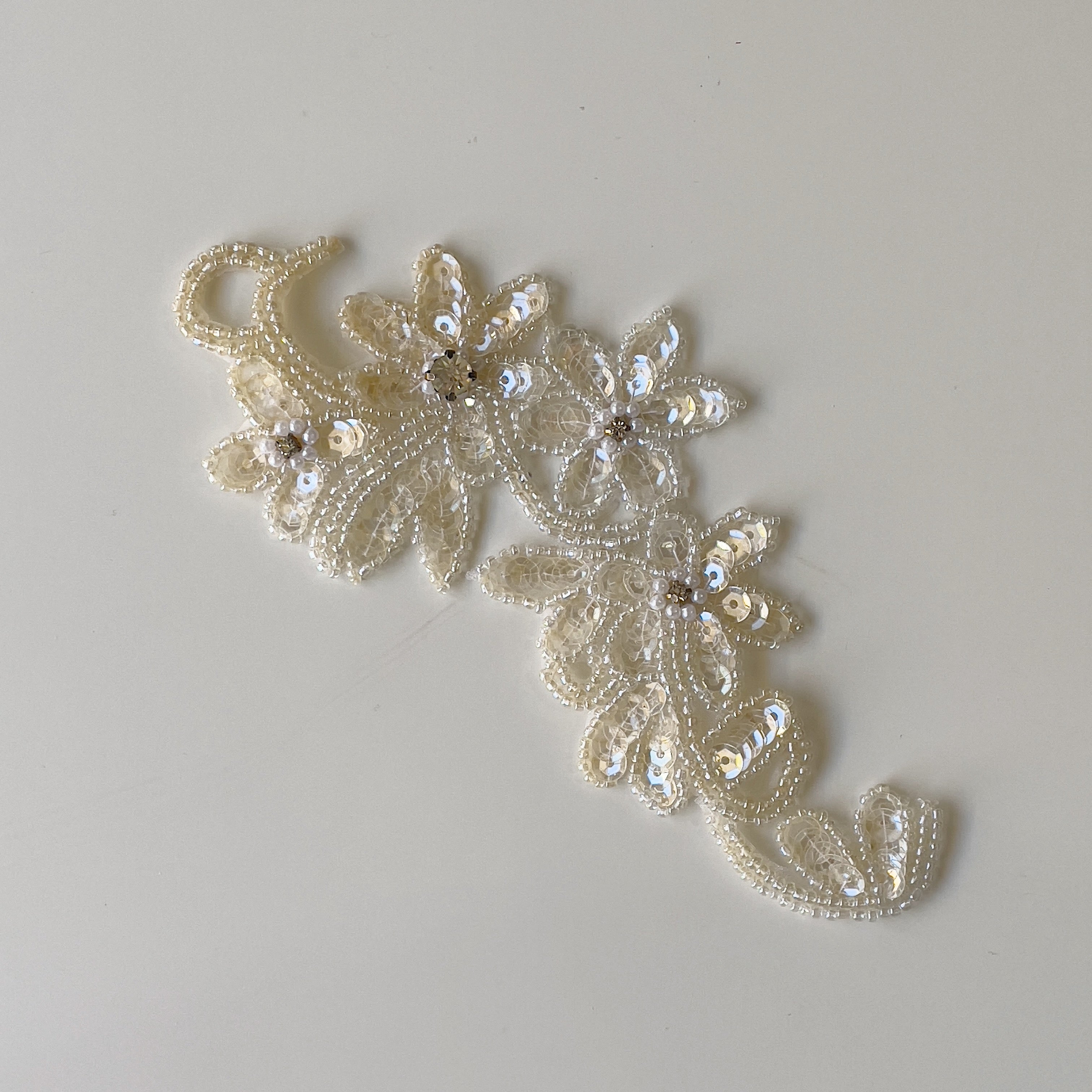 A sparkly single cream floral applique embellished with sequins and beads.  The flowers centres have a rhinestone decoration circled with tiny pearls.  The applique is laying flat on a white background.