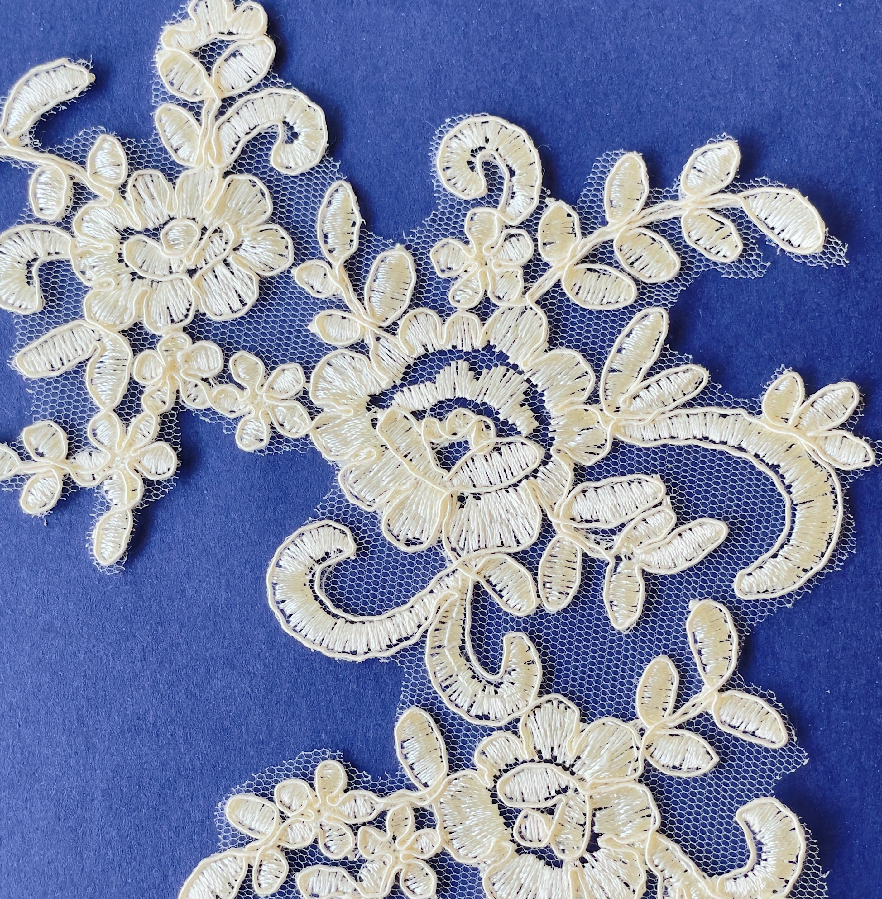 Close up view of cream embroidered and corded lace applique  with a floral design laying flat on a blue background.