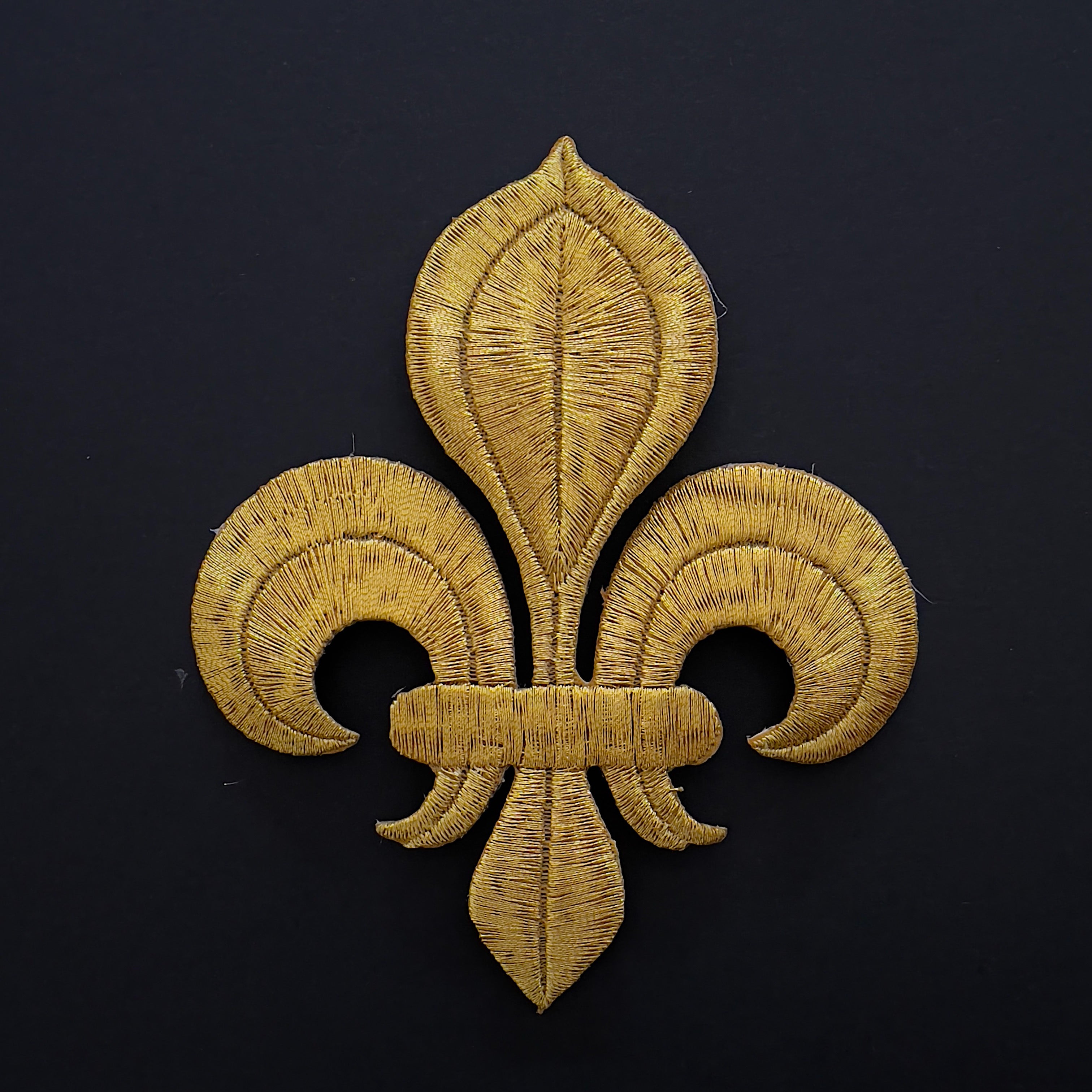 Gold fleur de lis applique embroidered with metallic thread laying flat on a black background.