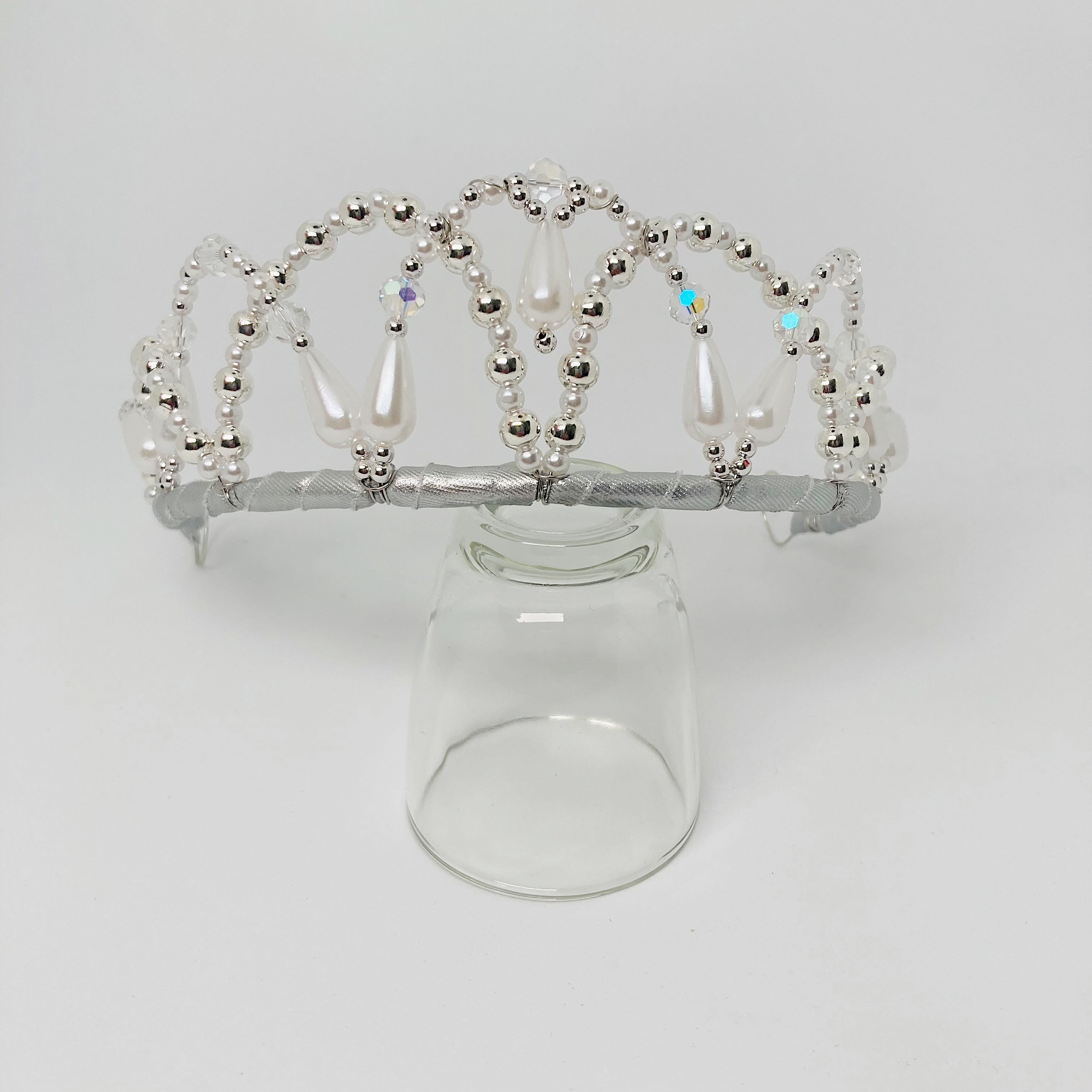 Front view of a tiara embellished with clear crystal, silver and frosted white beads.  The tiara is resting on a clear glass.  