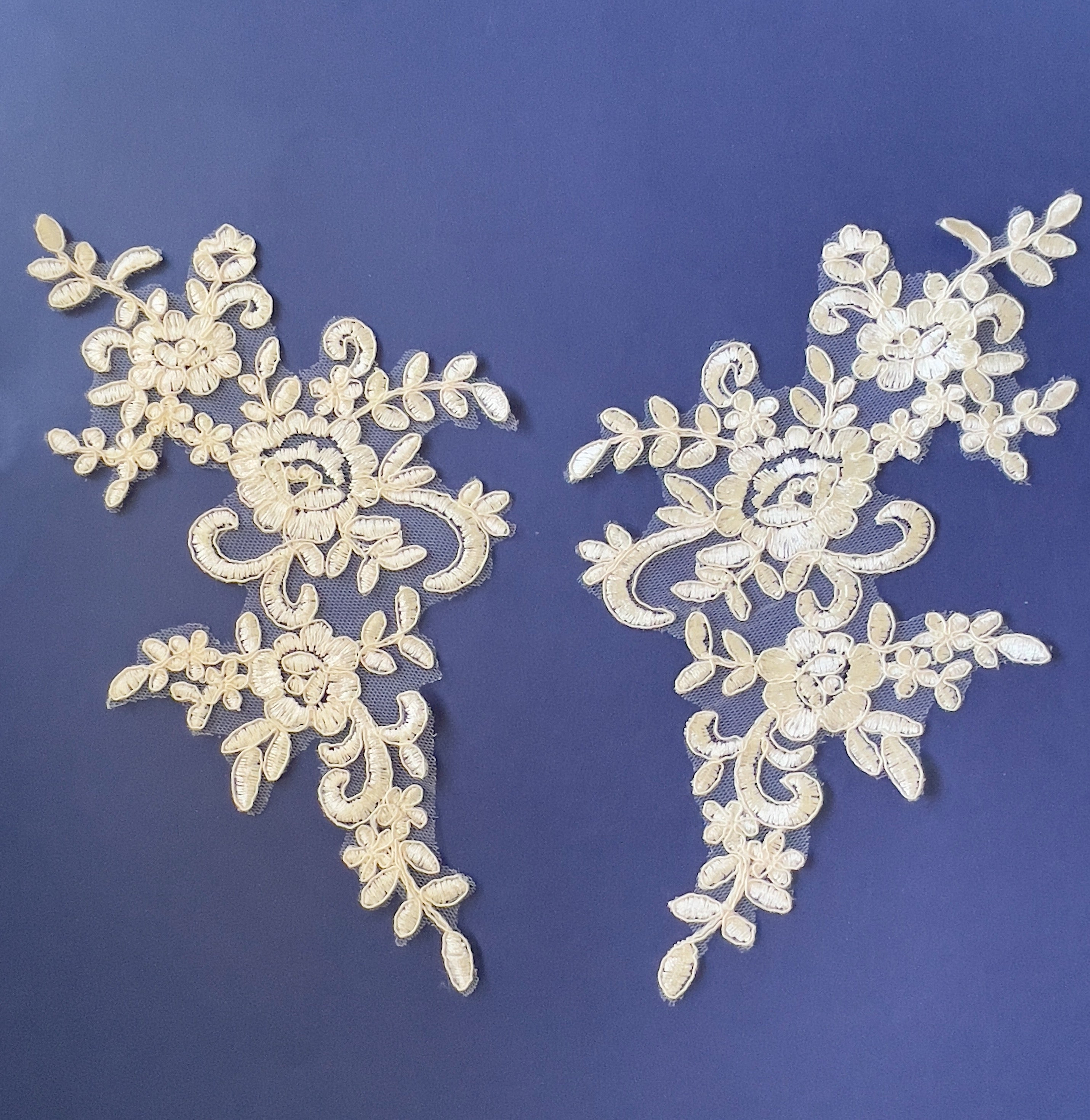 Cream embroidered corded floral applique pair laying flat on a blue background.